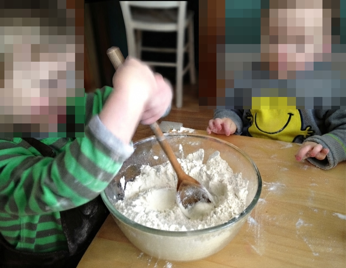 A young child in a green-and-grey striped top uses a wooden spoon to mix flour in a large glass bowl while another child watches.