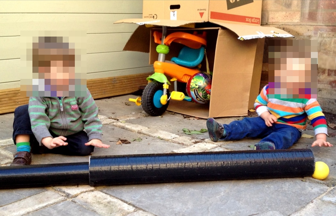 Two young children sitting on a patio playing with a long black plastic tube, watching a small yellow ball emerge from the end.