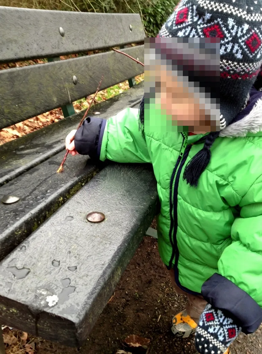 Child holding a stick to draw with rain water on a bench on a wet day.