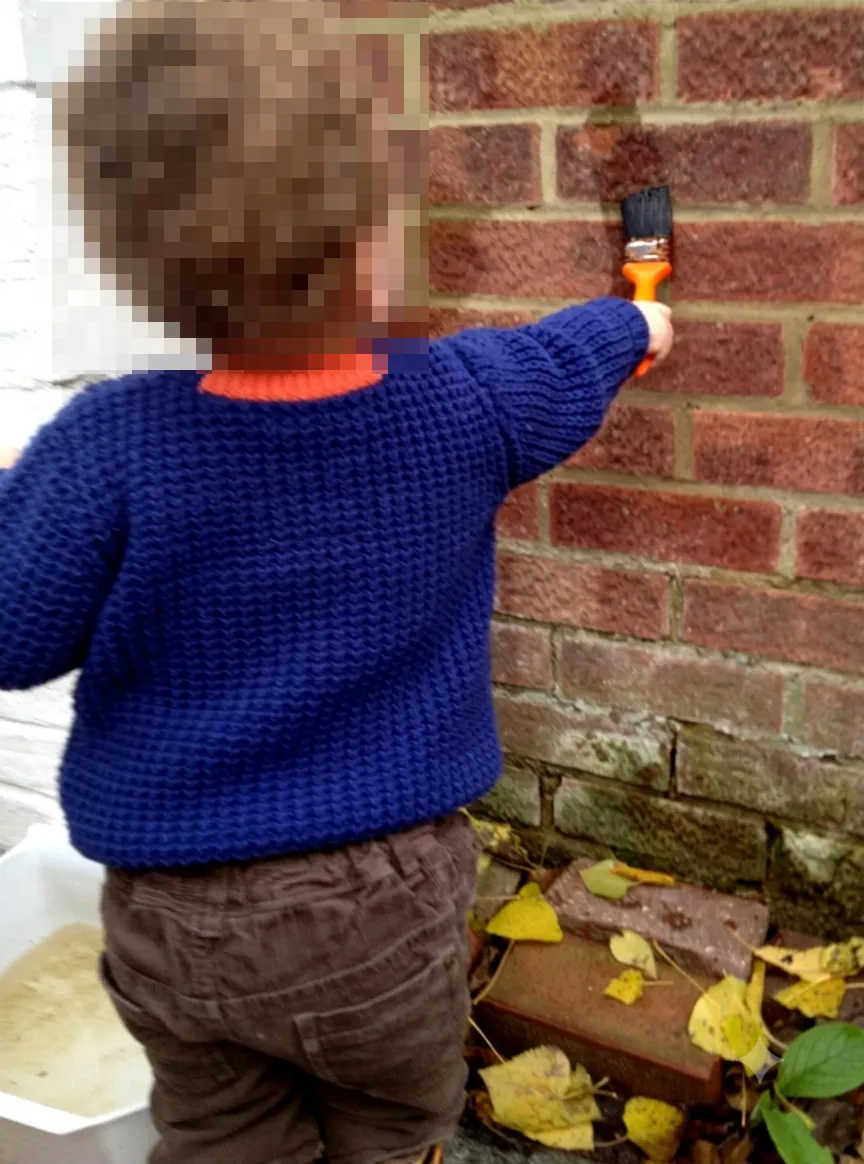 Child holding a paint brush and making marks on a brick wall using rain water from a pot beside him.