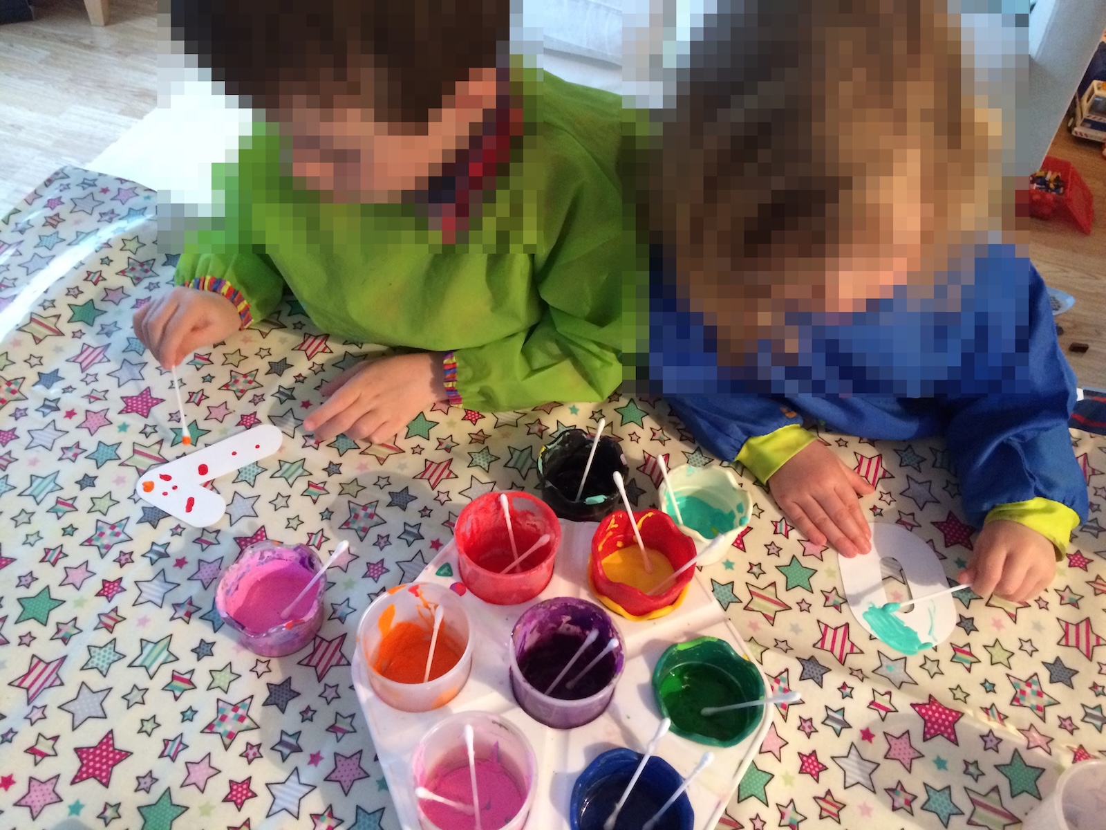 Two young children wearing art smocks sit at a star-patterned table, using cotton buds to paint cutout paper letters with colorful paints.
