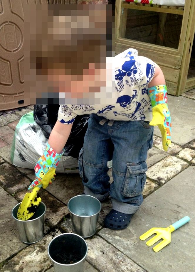 A young child in blue jeans and colourful gardening gloves using a yellow hand trowel to scoop soil into small metal buckets on a patio.