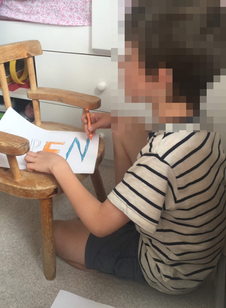 A young boy sitting on the floor and using an orange crayon to colour in a handmade 'OPEN' sign resting on a wooden chair.