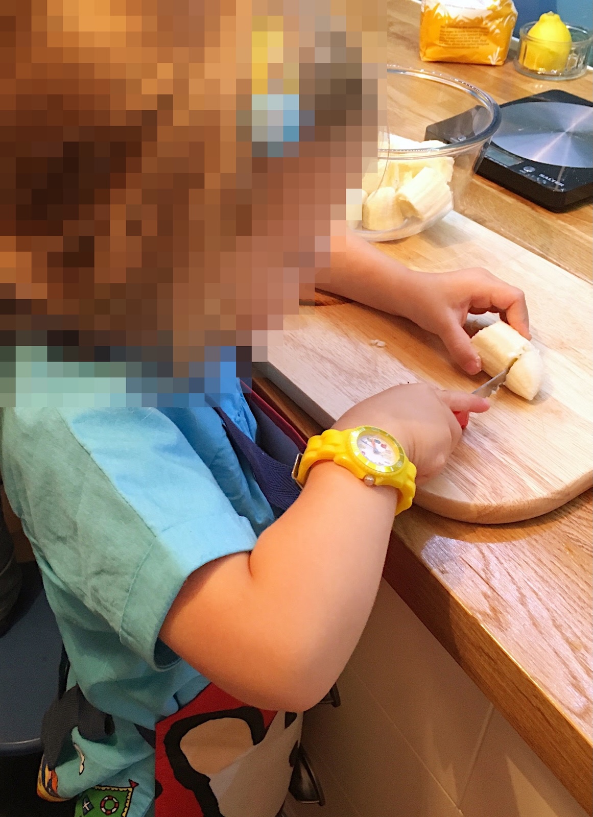 A young child wearing a yellow watch slicing a banana on a wooden chopping board on a kitchen counter.