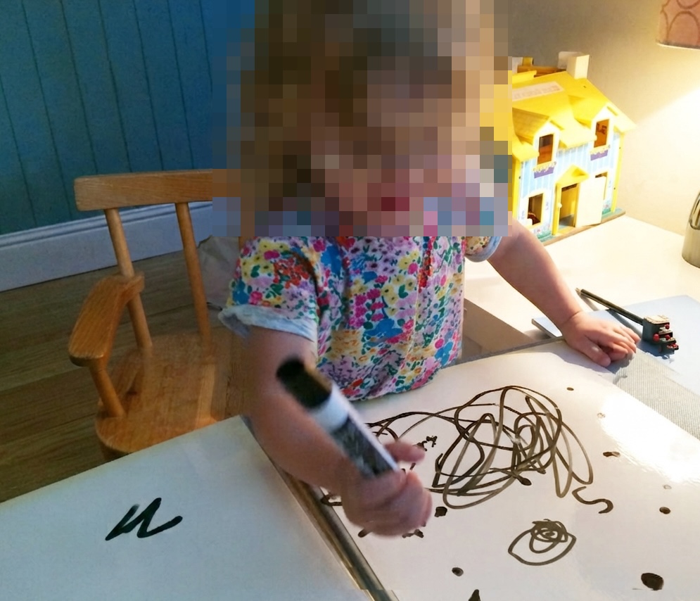 A young child in a floral shirt drawing dark scribbles on a white dry-erase board with a black marker.