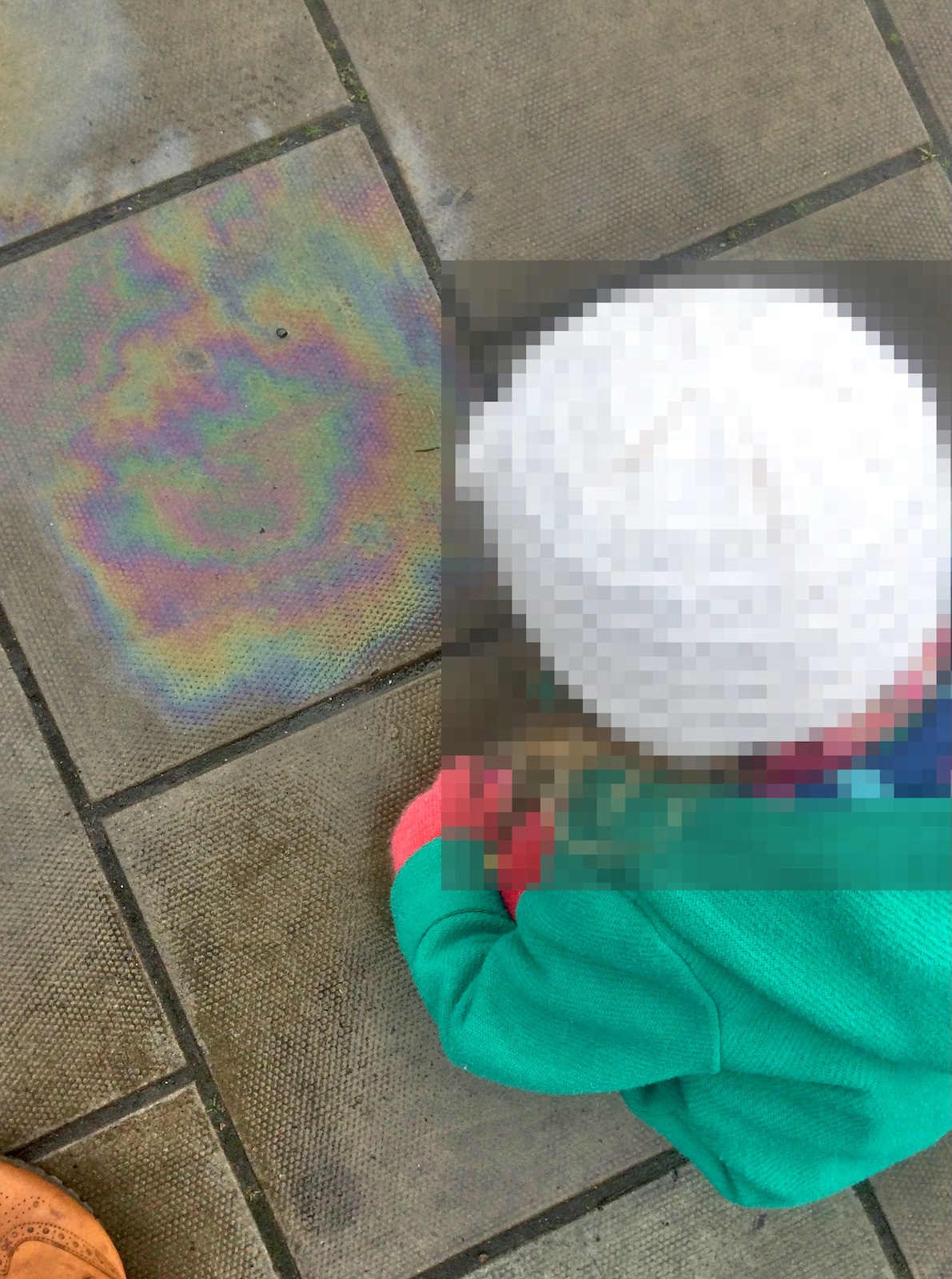 Overhead view of a young child in a green jacket crouching next to an iridescent rainbow oil slick on grey paving slabs.