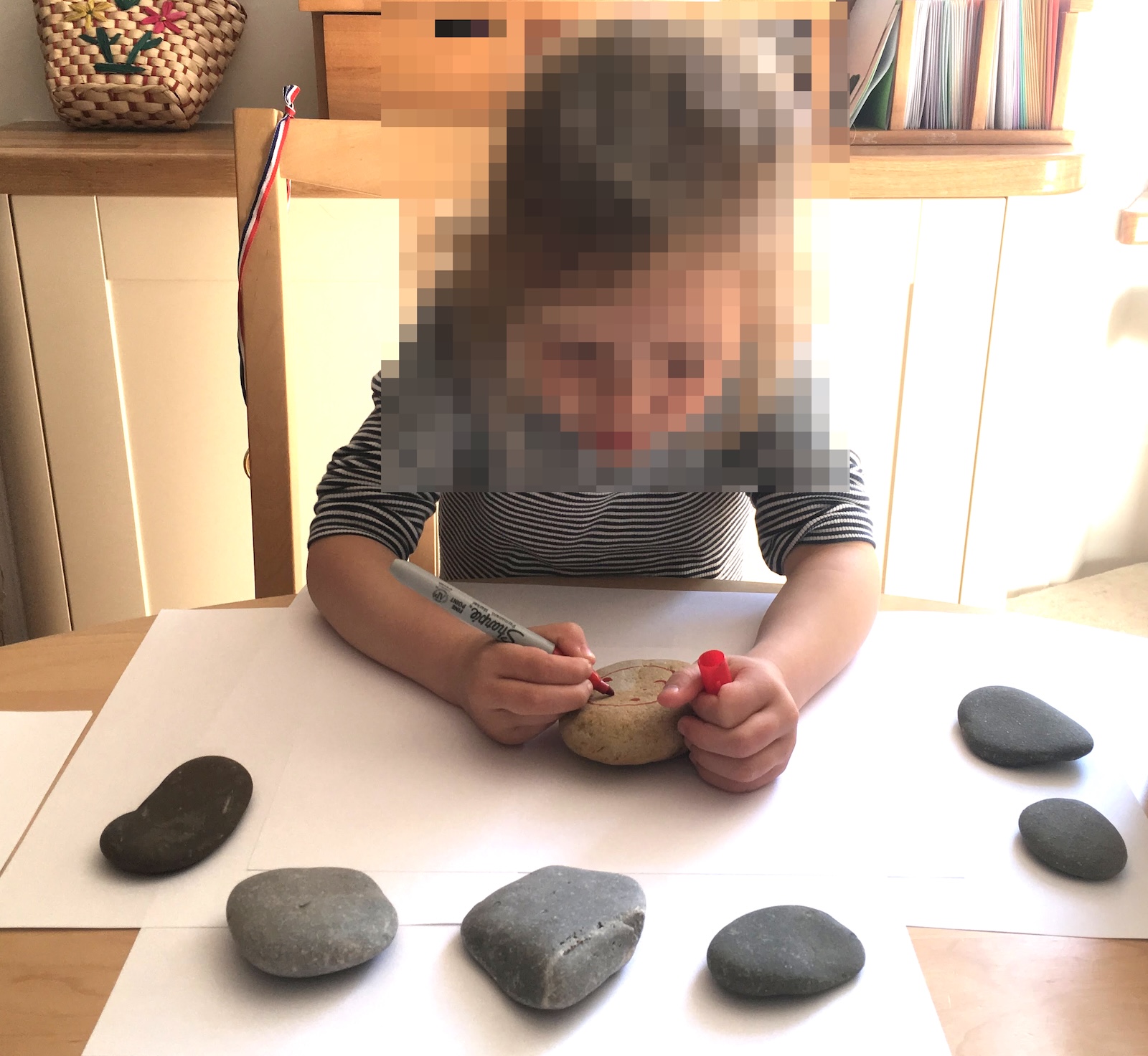 A young child sitting at a wooden table using a marker to decorate smooth stones placed on sheets of white paper.